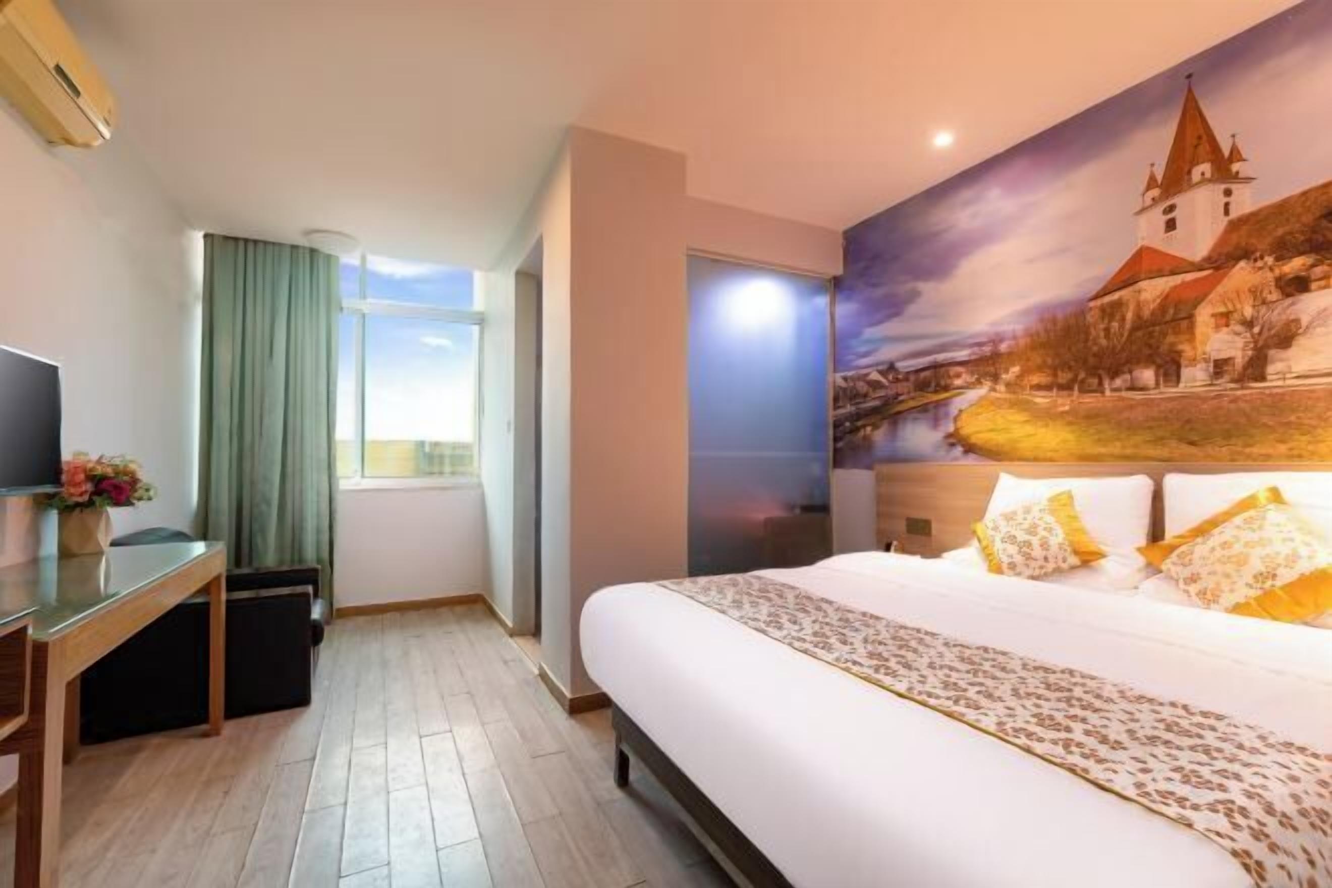 A hotel room with a large bed, a desk with a computer, a window with curtains, and a mural of a town on the wall.