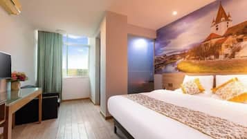 A hotel room with a large bed, a desk with a computer, a window with curtains, and a mural of a town on the wall.