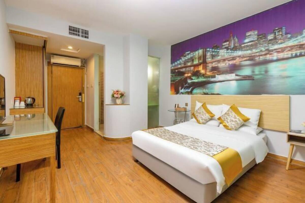 Quarto de hotel com cama, escrivaninha, cadeira, mesa de cabeceira, televisão na parede e pintura com vista urbana.