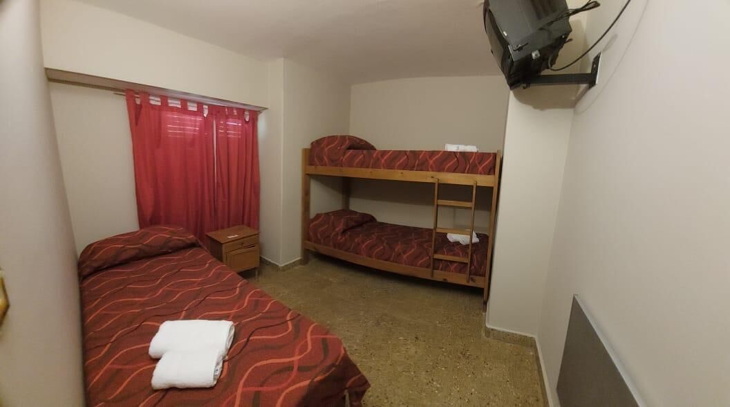A small, single-room hotel room with a bunk bed, a desk, a chair, a television mounted on the wall, and a window with red curtains.