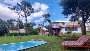 Un patio trasero con piscina, sillones de madera y un pequeño edificio de techo rojo.