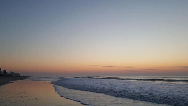 Una playa con mar tranquilo, cielo despejado y horizonte urbano a lo lejos.