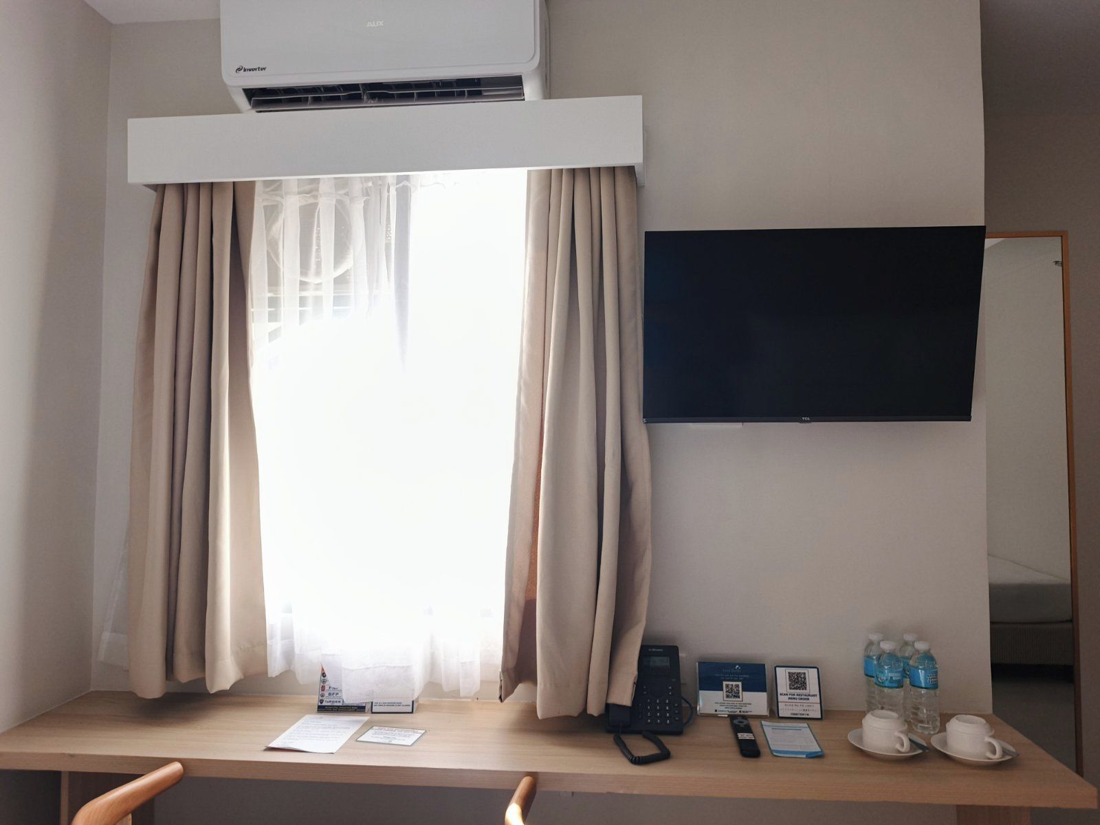 Habitación de hotel con escritorio, teléfono, pantalla digital, código QR y una botella de agua.