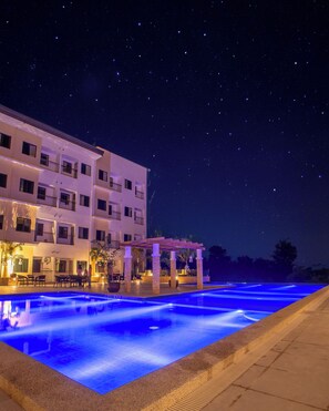 A nighttime view of a hotel with a swimming pool illuminated by blue lights, a covered outdoor seating area, and a building with multiple balconies. -  Panglao Vista Suites | FREE Airport & Seaport Pickup (Panglao)