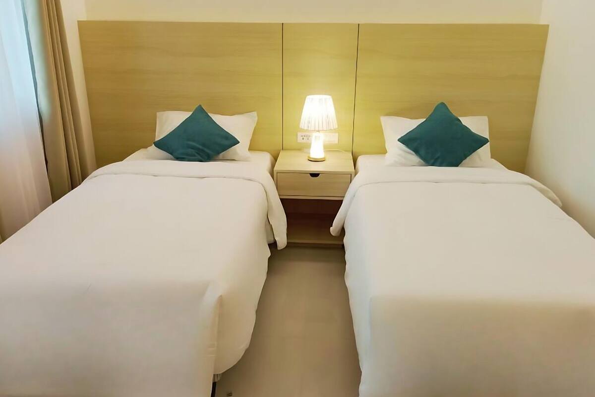 Una habitación de hotel con dos camas individuales, cada una con un almohadón verde azulado, una mesita de noche con una lámpara y un cabecero de madera.