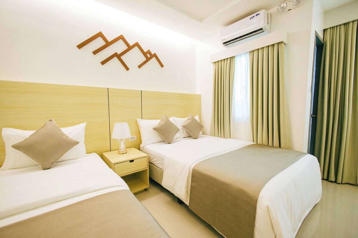 Habitación de hotel con dos camas, cabecera de madera, mesita de noche con lámpara y ventana con cortinas.