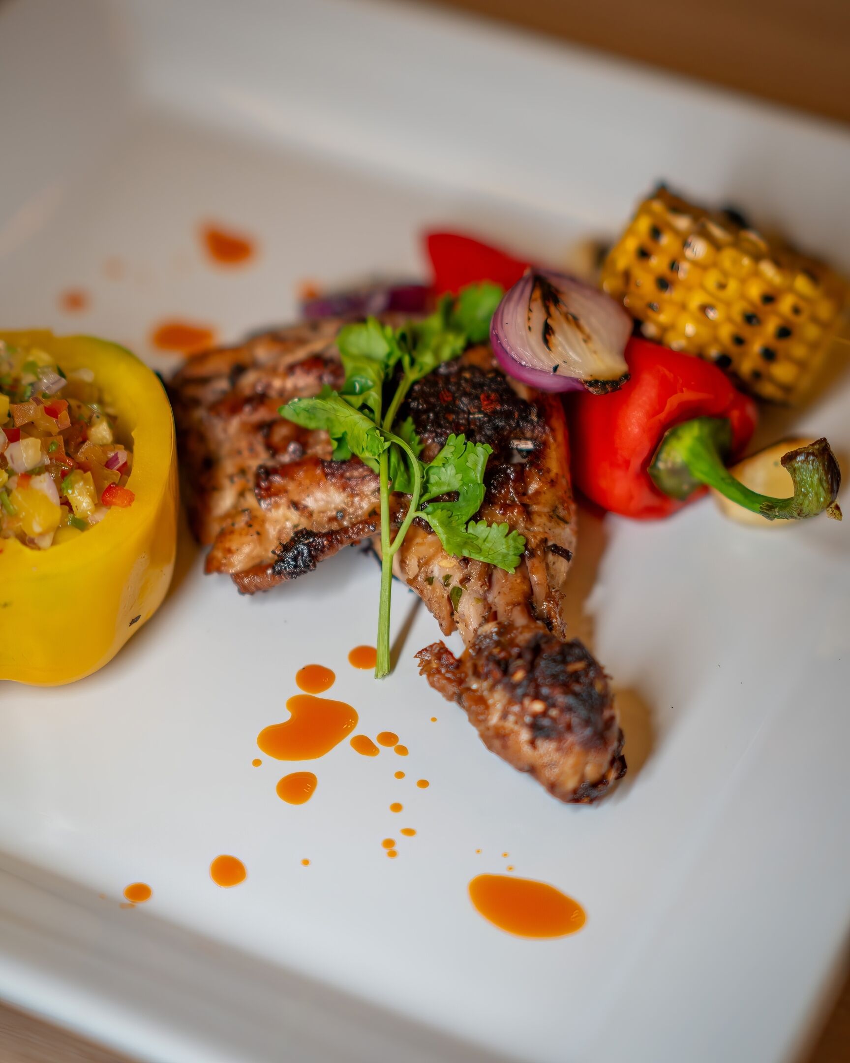 A plate with grilled chicken, a yellow bell pepper, a red bell pepper, corn on the cob, and a small salsa garnish.