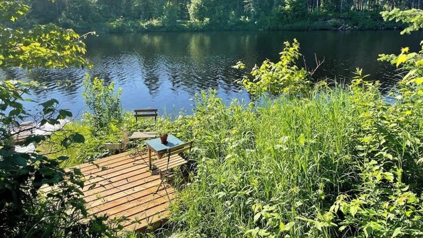 A wooden deck with a table and chairs overlooking a calm lake, surrounded by dense forest. - Spa, Relaxation, Forest & River | Le Marmontin (sainte-christine-d'auvergne)