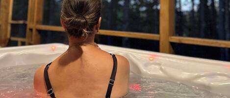 A person in a hot tub with a view of a forest through large windows.