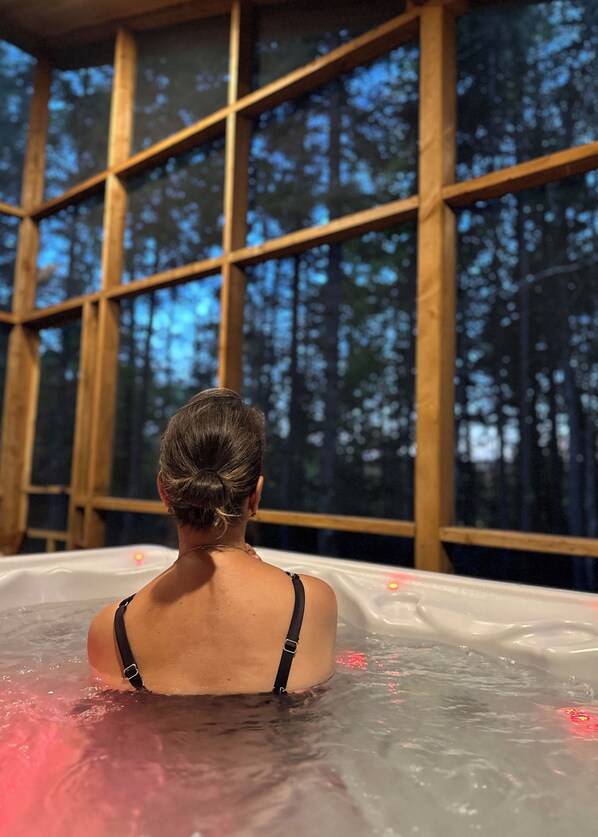 Une personne se détend dans un bain à remous, avec vue sur une forêt à travers de grandes fenêtres.