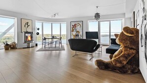 Un salon spacieux avec une grande fenêtre, une cheminée, un fauteuil noir et un grand ours en peluche.