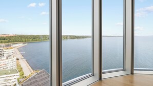 Une chambre d’hôtel située dans un immeuble de grande hauteur, dotée de grandes fenêtres du sol au plafond, offrant une vue sur une étendue d’eau calme et une côte lointaine.