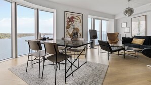 Un vaste espace de vie comprenant une table en verre, des chaises modernes, un canapé noir et une grande fenêtre donnant sur un plan d’eau.