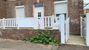 Ein Ziegelhaus mit zwei Fenstern, einem weißen Zaun und einem kleinen Garten mit rosa Blumen.