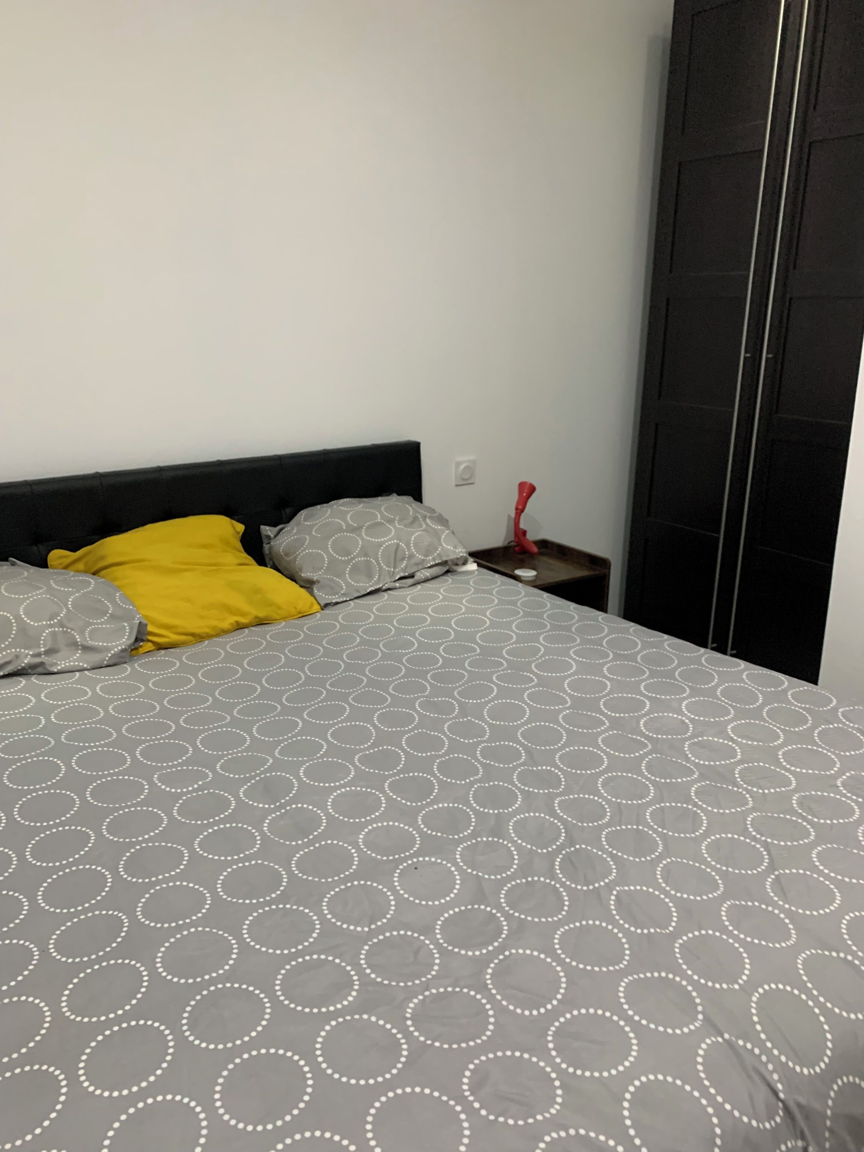 A bed with a grey and white circular pattern bedspread, a yellow pillow, and a grey pillow. There is a small wooden nightstand with a red object on it.