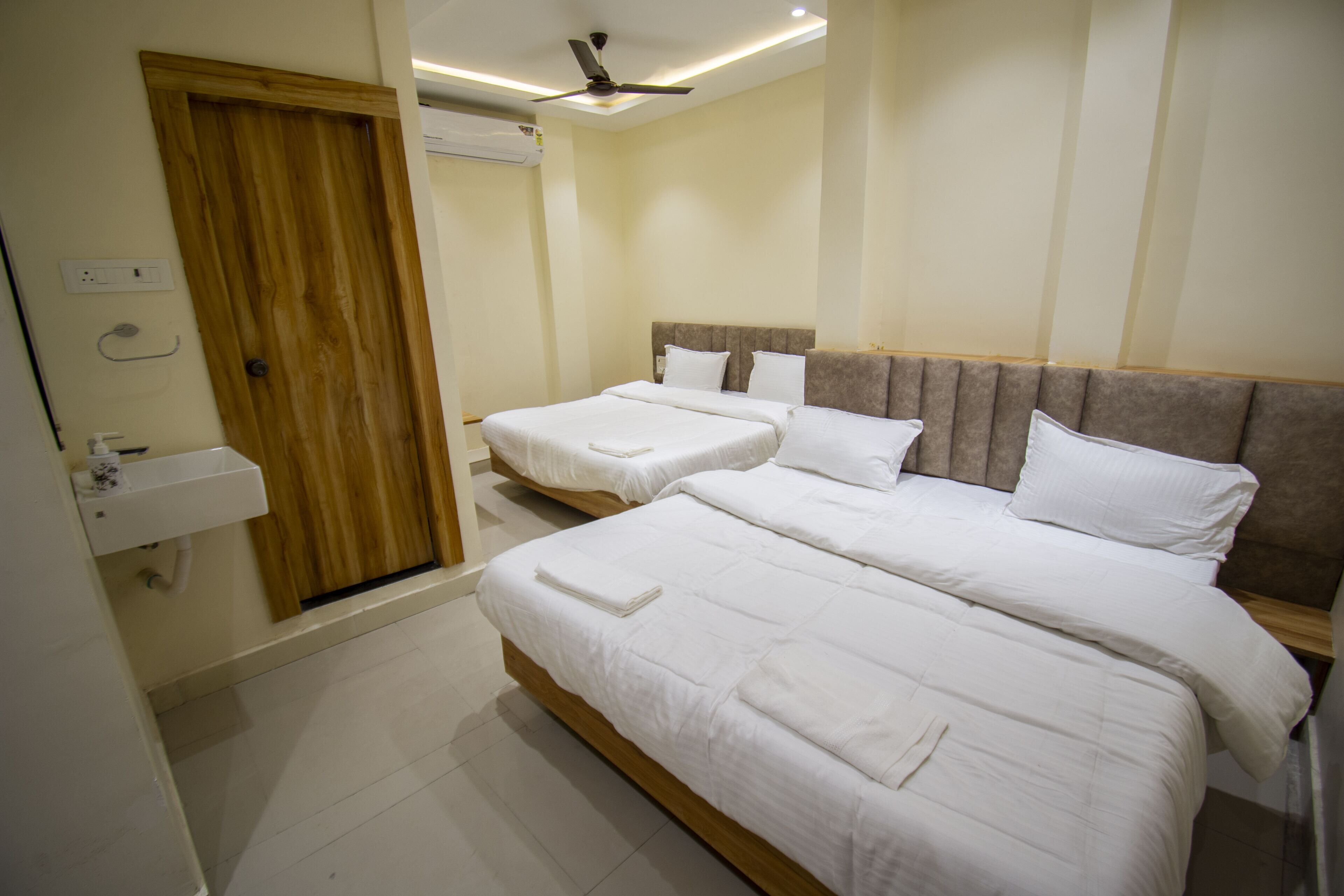 A hotel room with two beds, a wooden door, a ceiling fan, and a small sink.