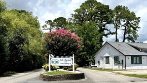 A sign for Bay Tree Golf & Racquet and Golf Colony IX, a white building with a sloped roof, and a large tree with pink flowers in front. - Awesome Studio at Bay Tree, 1st floor (Little River)