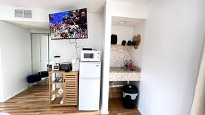 A compact kitchen with a microwave, toaster, and blender on a wooden shelf, a small refrigerator, and a TV mounted on the wall displaying an underwater scene. - LR2 Bay Tree great studio + pool in Little River (Little River)