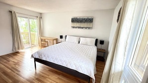A bedroom with a large bed, a desk, and a chair. There is a window with curtains, a painting on the wall, and a wooden floor. - LR2 Bay Tree great studio + pool in Little River (Little River)
