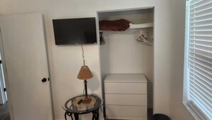 Una habitación compacta con una televisión montada en la pared, una mesita con lámpara, un aparador blanco y un armario con perchero.