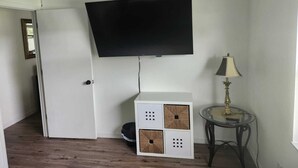 Una habitación con un televisor de pantalla plana montado en la pared, una mesita con una lámpara y un mueble blanco con cestas tejidas.