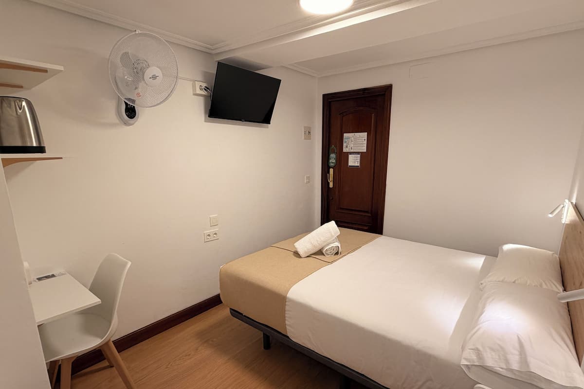 Une chambre d’hôtel avec un lit, un bureau, une chaise, un téléviseur fixé au mur et une porte en bois.