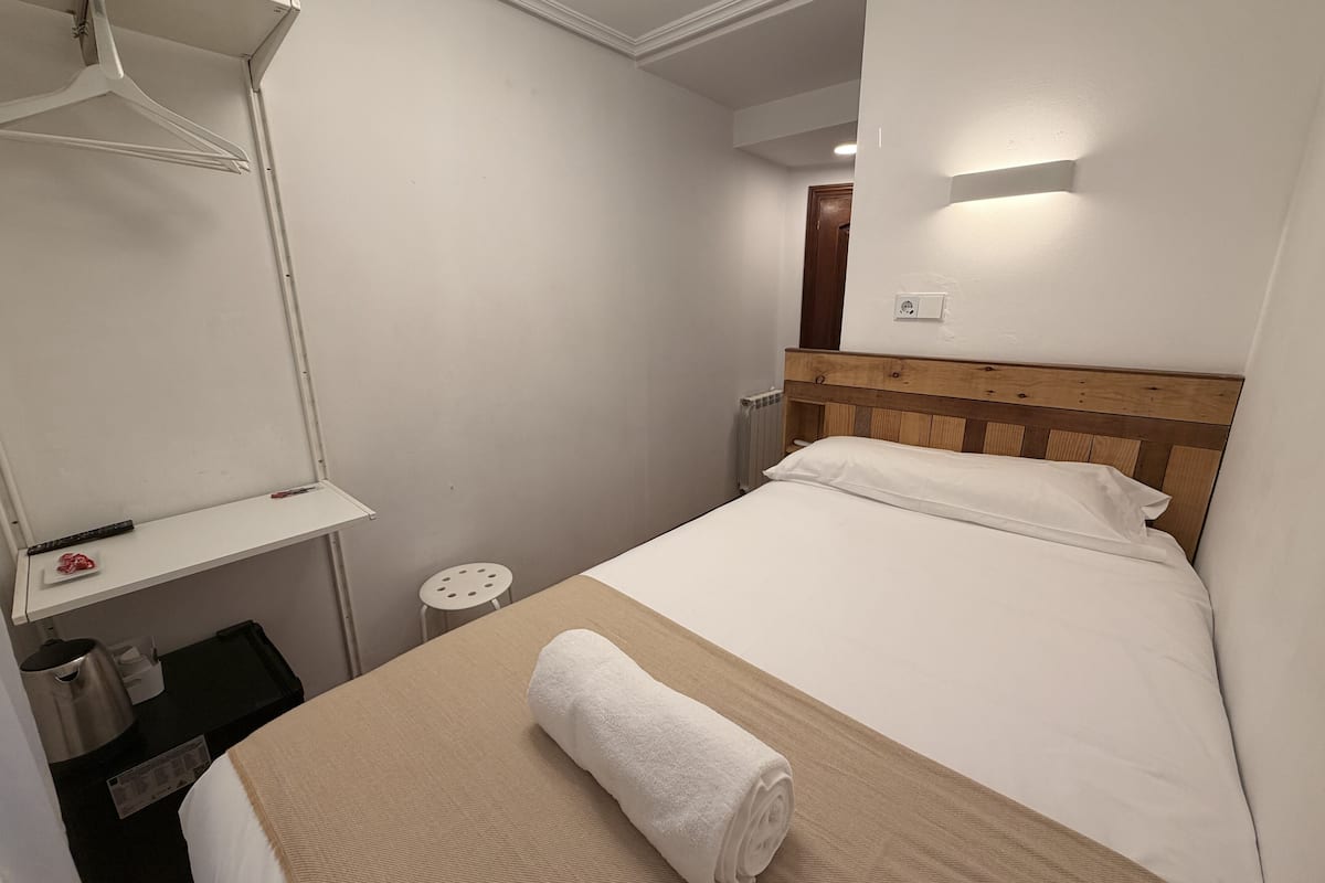 Une chambre d’hôtel de taille réduite, avec un lit simple, une tête de lit en bois, une petite table de chevet, une serviette blanche sur le lit et une étagère fixée au mur.