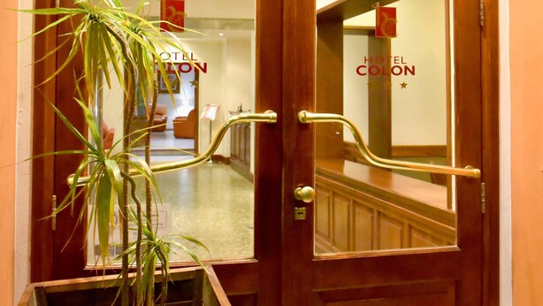 Une entrée d’hôtel en bois, agrémentée d’un pot de fleurs, d’un miroir et d’une enseigne indiquant « HÔTEL COLON ».