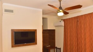 Une pièce dotée d’un ventilateur de plafond, d’un téléviseur fixé au mur, d’une chaise en bois et d’une fenêtre avec des rideaux.