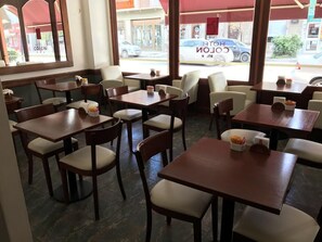 A hotel restaurant with multiple tables and chairs, large windows with a view of the street, and a sign reading "HOTEL COLON".