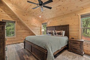 Quarto com teto inclinado, cama com manta acolchoada, mesa de cabeceira e janela com vista para a floresta.