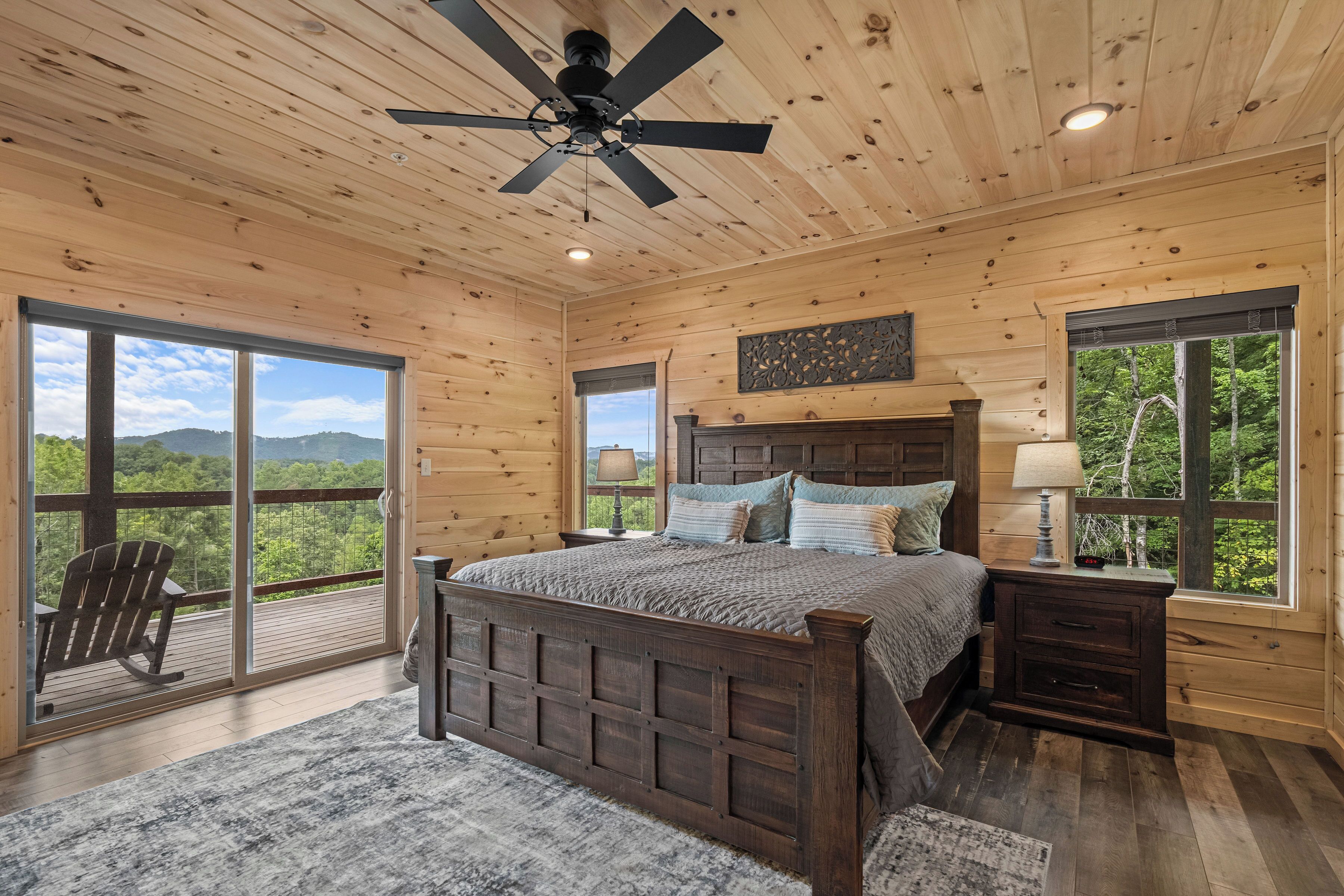 A wooden cabin bedroom with a large bed, two nightstands, a ceiling fan, and a view of the forest through large windows.