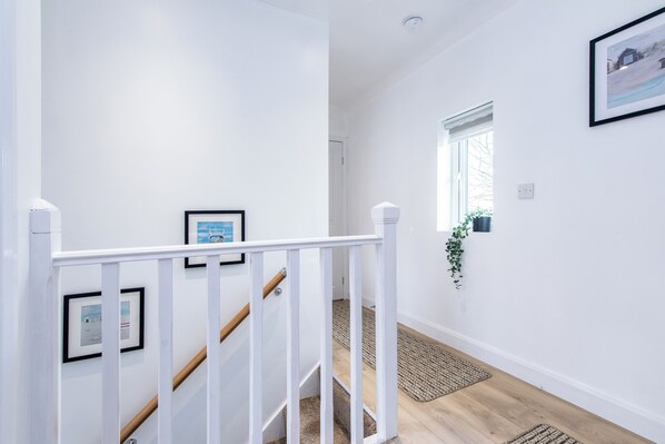 A clean, white-walled hallway with a wooden staircase, framed pictures, a window with a view, and a potted plant.