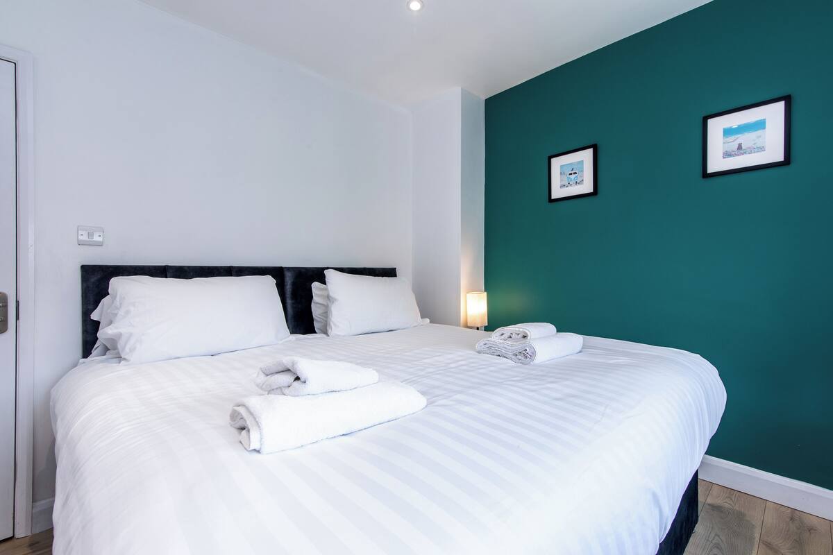 A neatly made bed with white linens and towels, a dark headboard, and teal walls with framed pictures.