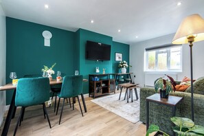 A modern living room with teal walls, a wooden dining table, teal chairs, a sofa, a TV, and a floor lamp.