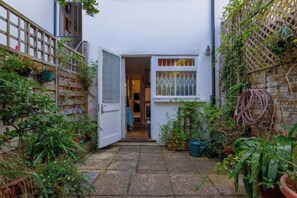A narrow courtyard with a white door leading to a room, surrounded by potted plants and a wooden fence. - Delightful 3BD Family Home w Garden, Finsbury Park (Greater London)
