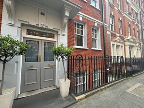 A row of red brick townhouses with white window frames, a grey door with a brass handle, and a black wrought iron fence. - Superbly Located 1BD Flat, Heart of Fitzrovia (Greater London)