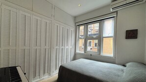 A bedroom with a bed, a piano, and a window with a view of buildings. - Superbly Located 1BD Flat, Heart of Fitzrovia (Greater London)
