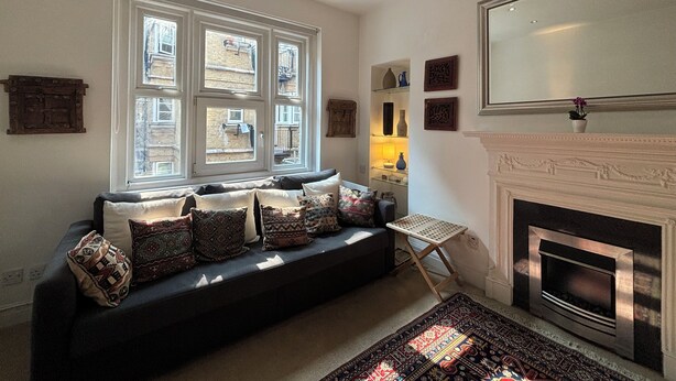 A living room with a dark-colored sofa, patterned cushions, a small side table, a fireplace, and a large window with multiple panes. - Superbly Located 1BD Flat, Heart of Fitzrovia (Greater London)