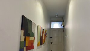 A hallway with three abstract paintings on the wall, a white door at the end, and a small window with a view of the sky. - Superbly Located 1BD Flat, Heart of Fitzrovia (Greater London)