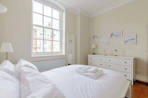 A bedroom with a large window, a bed with white linens, a dresser with drawers, and wall-mounted artwork.