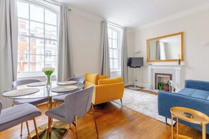 A living room with a dining table, chairs, a sofa, a blue armchair, a fireplace, and a large mirror.