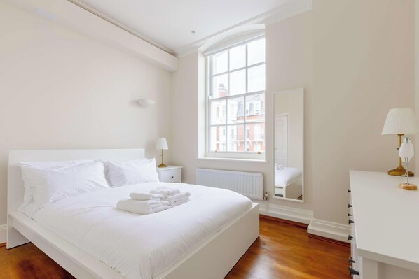 A white bedroom with a large window, a bed with white linens, a bedside table with a lamp, and a dresser with a lamp.