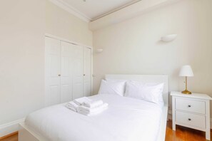 Un lit bien fait, avec du linge de lit blanc et une serviette pliée, une table de chevet blanche avec une lampe, et une armoire blanche.