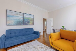 Uma sala de estar com um sofá azul, uma poltrona amarela e duas pinturas abstratas na parede.