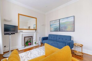 A living room with a blue sofa, a yellow armchair, a fireplace, a TV, and artwork on the wall.