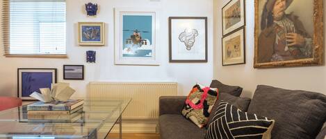 A living room with a glass coffee table, a sofa with decorative pillows, and various framed artworks on the walls.