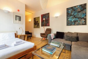 A modern hotel room with a bed, a sofa, a coffee table with books, a chair, and abstract wall art. - One of a Kind 2BDR Flat in Shoreditch (London)