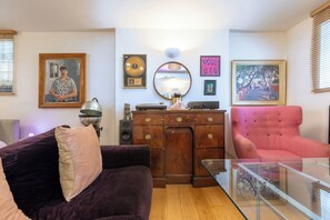 A living room with a dark-colored sofa, a glass coffee table, a wooden cabinet, and a pink armchair. There are framed pictures on the walls and a mirror above the cabinet. - One of a Kind 2BDR Flat in Shoreditch (London)