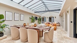 Une salle à manger spacieuse avec un haut plafond et des panneaux de verre, une longue table dressée pour le repas et des sièges confortables.